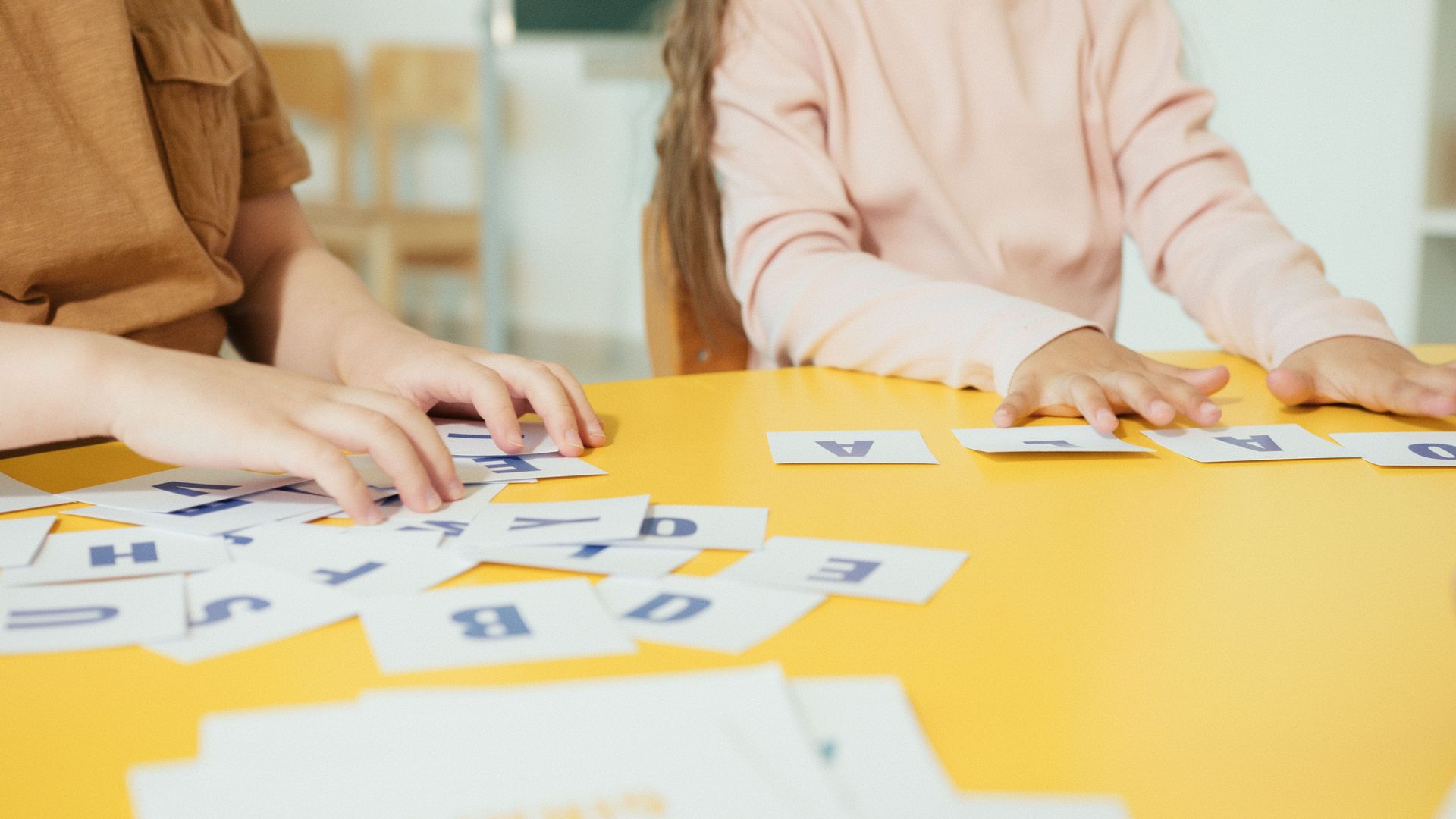 Children arranging alphabet cards on a yellow table in a classroom setting.