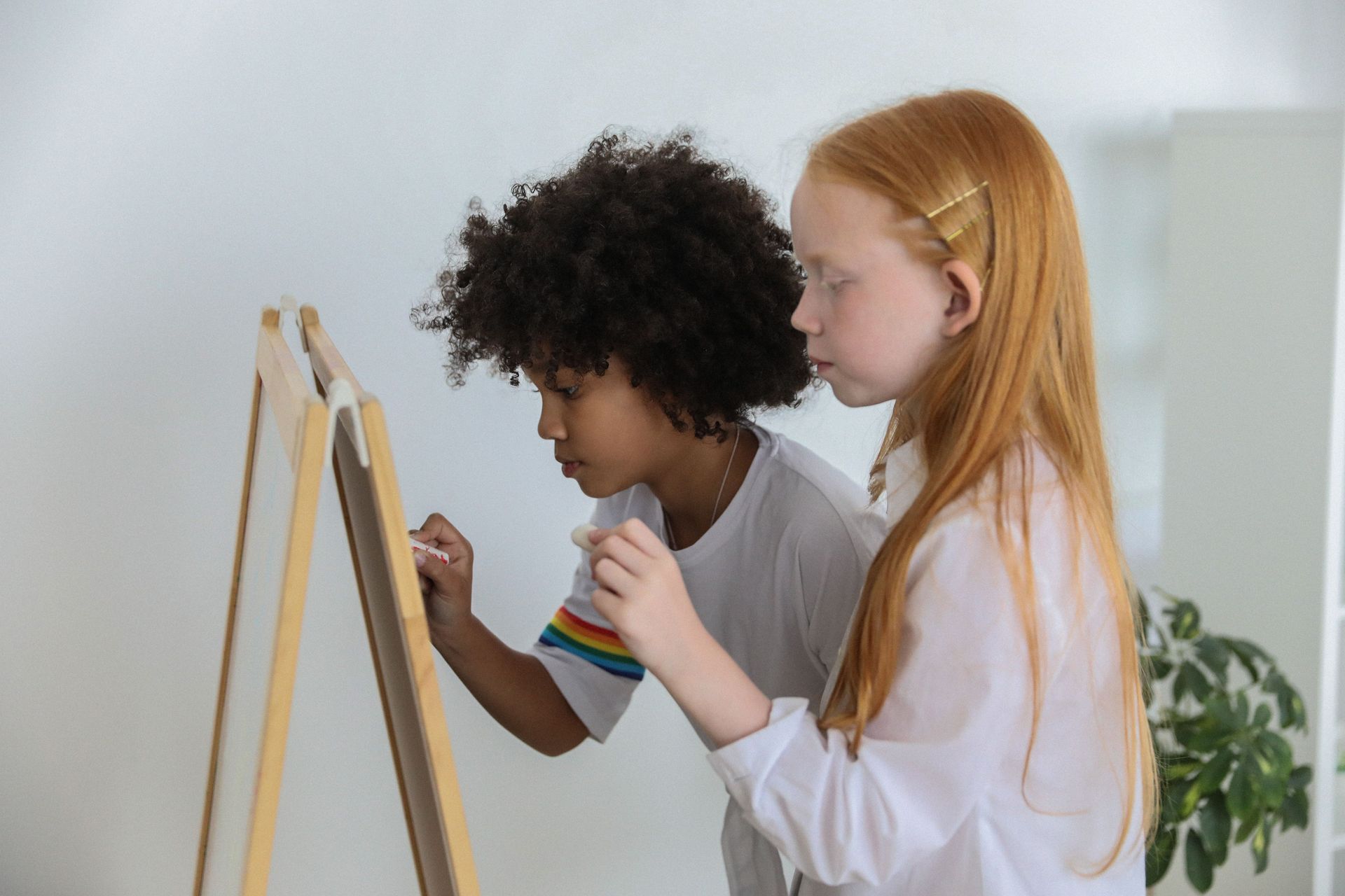 Side view of focused diverse kids standing and drawing on easel chalkboard in light room